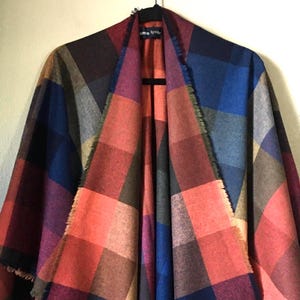May include: A large, open-front poncho or wrap featuring a bold, multi-colored plaid pattern. The fabric displays a grid of deep blue, vibrant red, muted olive green, and purple squares. The edges are finished with a raw, frayed fringe detail.