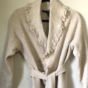 May include: A beige knit cardigan with a shawl collar and fringe trim. The long-sleeved sweater has a matching belt that ties at the waist. The cardigan is hanging on a black hanger against a white wall.