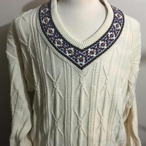 May include: Cream-colored V-neck sweater with a cable knit design. The neckline features a patterned border with blue, brown, and white details. The sweater has long sleeves and a ribbed hem and cuffs. A classic, comfortable knitwear piece.