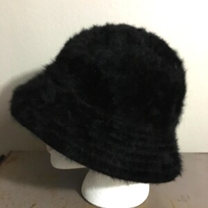 Op de afbeelding: Een zwarte, donzige vissershoed. De hoed heeft een afgeronde kroon en een iets naar beneden hellende rand. De hoed is gemaakt van een zacht, getextureerd materiaal en wordt getoond op een witte mannequinhoofd.