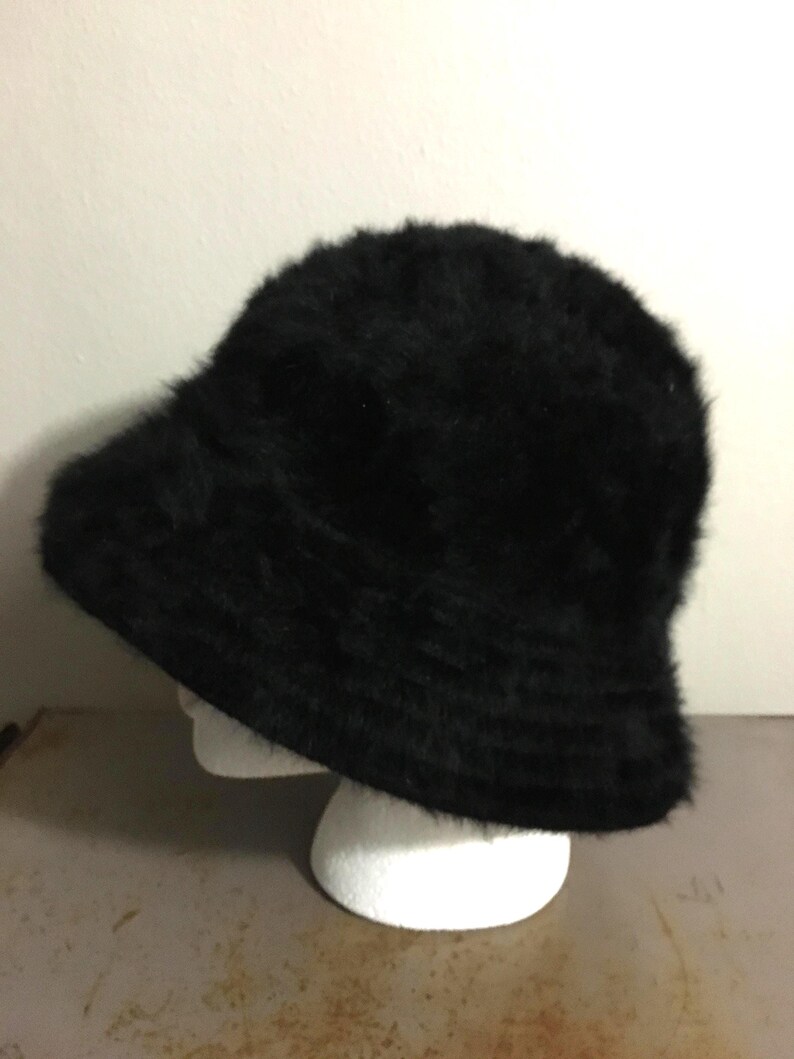 Op de afbeelding: Een zwarte, donzige vissershoed. De hoed heeft een afgeronde kroon en een iets naar beneden hellende rand. De hoed is gemaakt van een zacht, getextureerd materiaal en wordt getoond op een witte mannequinhoofd.