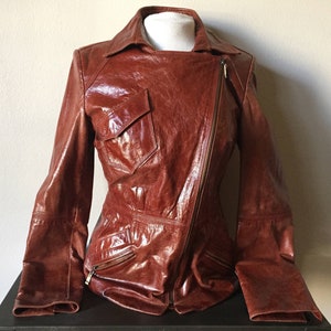 May include: A brown leather jacket with a zipper closure and a large pocket on the left side. The jacket has a slightly asymmetrical design.