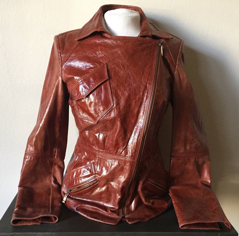 May include: A brown leather jacket with a zipper closure and a large pocket on the left side. The jacket has a slightly asymmetrical design.
