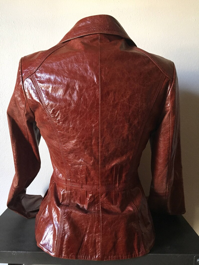 May include: A brown leather jacket with a collared neckline and long sleeves. The jacket has a fitted silhouette and a smooth, glossy finish.