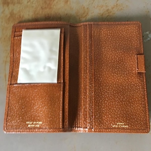 May include: A tan textured bifold wallet featuring a clear plastic ID window on the left side, multiple card slots, and gold embossed text on the interior that reads "GENUINE LEATHER" and "MADE IN CANADA" on the right side panel.