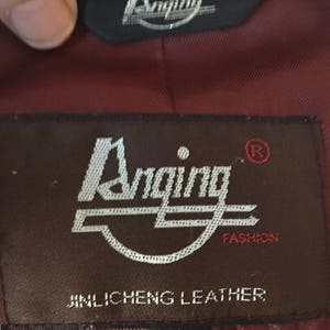 May include: A brown leather jacket label with the text "Anging Fashion" and "Jinlichen Leather" in white lettering. The label also has a registered trademark symbol and the letter "L" for size.