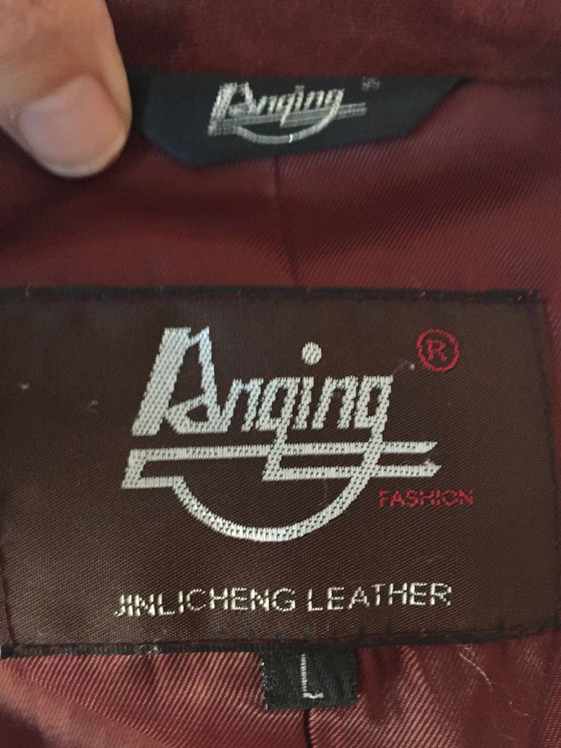 May include: A brown leather jacket label with the text "Anging Fashion" and "Jinlichen Leather" in white lettering. The label also has a registered trademark symbol and the letter "L" for size.