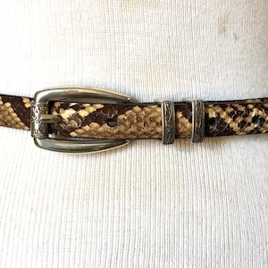 May include: A slim belt featuring a brown and tan snakeskin pattern with a polished silver-tone metal buckle and two matching silver-tone metal keepers. The belt is secured around a white mannequin torso, showcasing the textured scale design.