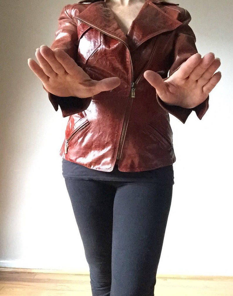 May include: A brown leather jacket with a zipper closure and a collar. The jacket is worn by a person with their hands raised in front of them.