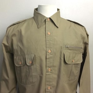 May include: A long-sleeved tan button-down shirt featuring a pointed collar, shoulder epaulets, and multiple front pockets. The shirt includes two lower patch pockets with buttoned flaps and an additional zippered pocket above the left chest pocket.