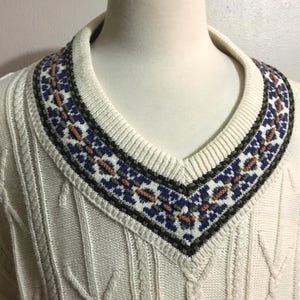 May include: An off-white V-neck sweater featuring a textured cable knit pattern throughout. The neckline is accented with a decorative band displaying a geometric pattern in shades of dark blue, grey, and muted orange against a white background.