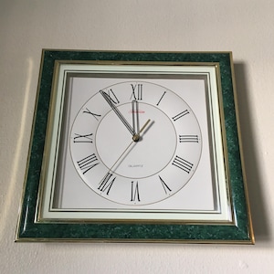May include: Square quartz clock with a green marble-look frame and gold trim. The clock face is white with black Roman numerals and hands. The word "QUARTZ" is printed at the bottom of the clock face.