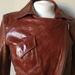 May include: A brown leather jacket with a zipper closure and a large pocket on the front. The jacket has a shiny finish and a classic design.