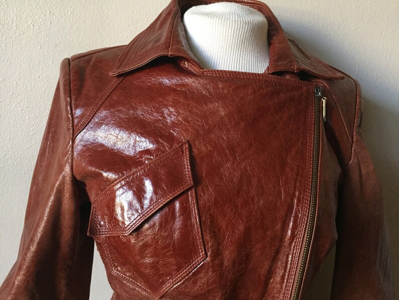 May include: A brown leather jacket with a zipper closure and a large pocket on the front. The jacket has a shiny finish and a classic design.