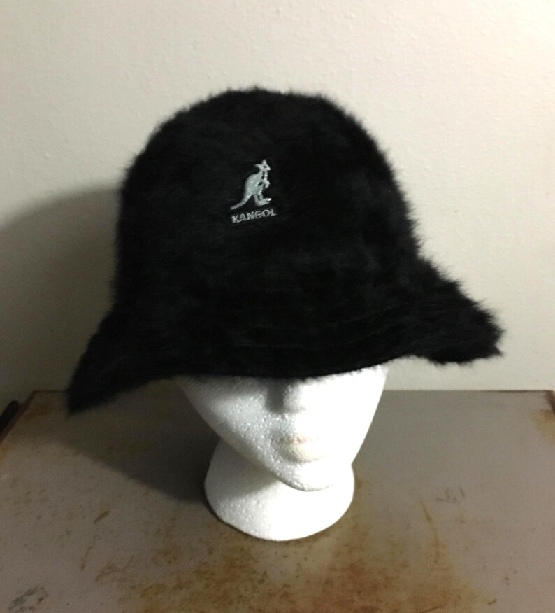 Op de afbeelding: Een zwarte Kangol-emmerhoed met een zachte, donzige textuur. De hoed heeft een klein, wit geborduurd kangoeroelogo. De hoed wordt gepresenteerd op een witte mannequinhoofd.