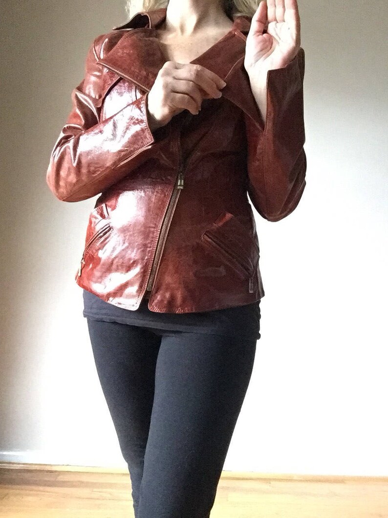 May include: A brown leather jacket with a zipper closure and a collar that can be worn up or down. The jacket has a fitted silhouette and is made of a shiny, glossy leather.