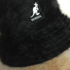 Op de afbeelding: Een zwarte vissershoed met een zachte, donzige textuur. De hoed heeft een witte patch met het woord "KANGOL" en een kangoeroelogo. Een modieus accessoire.