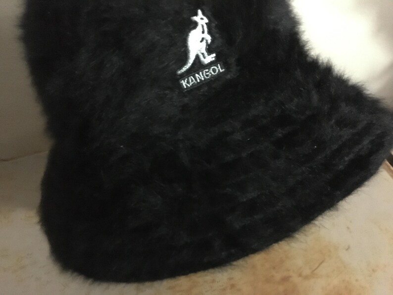 Op de afbeelding: Een zwarte vissershoed met een zachte, donzige textuur. De hoed heeft een witte patch met het woord "KANGOL" en een kangoeroelogo. Een modieus accessoire.
