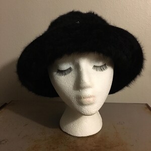 Op de afbeelding: Een zwarte bont hoed met een brede rand. De hoed wordt getoond op een witte mannequin hoofd. De hoed heeft een zacht, getextureerd uiterlijk en is waarschijnlijk gemaakt van imitatiebont. De rand is licht gebogen.