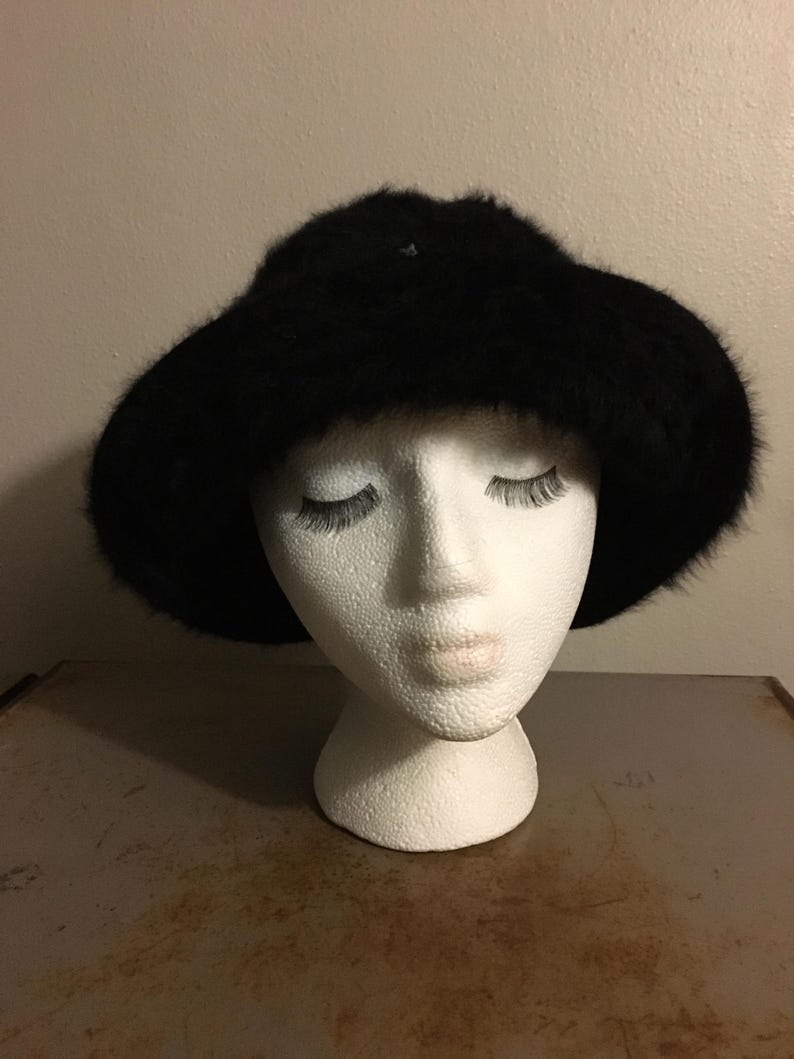 Op de afbeelding: Een zwarte bont hoed met een brede rand. De hoed wordt getoond op een witte mannequin hoofd. De hoed heeft een zacht, getextureerd uiterlijk en is waarschijnlijk gemaakt van imitatiebont. De rand is licht gebogen.