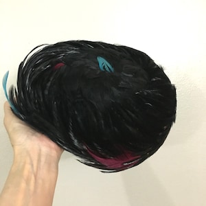 May include: A black feather hat with accents of teal and magenta. The hat is round and appears to be made of many individual feathers. The hat is being held by a person's hand.