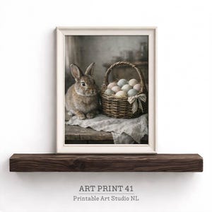 Op de afbeelding: Een ingelijste kunstdruk met een bruin konijn naast een gevlochten mand vol pastelkleurige eieren. De mand is vastgebonden met een strik. De print staat op een houten plank. De tekst "ART PRINT 41 Printable Art Studio NL" staat onderaan.