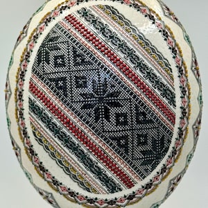 Op de afbeelding: Een ovaalvormig ei versierd met ingewikkelde, verhoogde was-resist patronen in zwart, rood, groen en geel. Het ontwerp bevat geometrische arceringen, stervormige motieven en delicate bloemenranden in symmetrische banden.