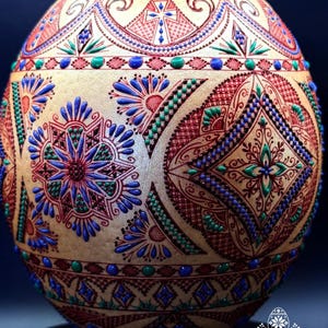 Può includere: Un guscio d'uovo di forma ovale decorato con intricati motivi geometrici e floreali dipinti a mano in rosso, blu e verde. La superficie presenta punti in rilievo testurizzati e linee sottili disposte in motivi simmetrici, inclusi rombi e stelle.