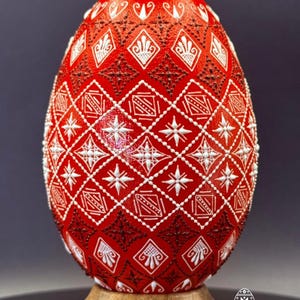 Może przedstawiać: Jajko w żywym czerwonym kolorze, ozdobione skomplikowanymi białymi i ciemnobrązowymi geometrycznymi wzorami, w tym rombami, gwiazdami i kreskowaniem. Jajko jest umieszczone na gładkiej, jasnej drewnianej podstawce, ukazując detale reliefu.