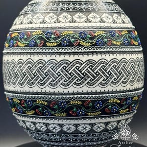 Könnte beinhalten: Ein dekoratives Ei mit komplexen schwarz-weißen geometrischen Mustern, darunter ein zentrales Flechtband und horizontale Reihen von Blumenmotiven in Rot, Blau, Grün und Gelb. Das Ei steht auf einem kleinen Holzsockel und zeigt den Text Atelier Zestrea.