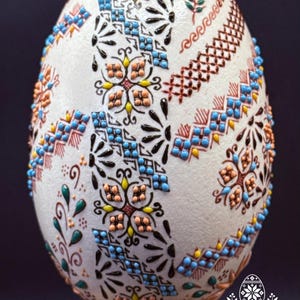 May include: A white egg decorated with intricate raised patterns in black, blue, peach, yellow, and green. The design features geometric shapes, floral motifs, and cross-hatching. The egg rests on a wooden base, with the text Atelier Zestrea visible.