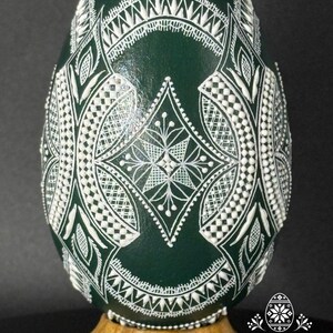 Könnte beinhalten: Ein dunkelgrünes Ei, verziert mit komplexen weißen Reliefmustern aus geometrischen Formen, Rautenmotiven und feinen Linien. Das Ei steht auf einem hellbraunen Holzsockel. Der Text Atelier Zestrea Anamaria Buraciuc ist zu sehen.