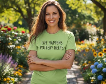 May include: A light green t-shirt with the text "LIFE HAPPENS POTTERY HELPS." The shirt is worn by a person standing in a garden with a variety of colorful flowers and trees. The person is smiling with their arms crossed.