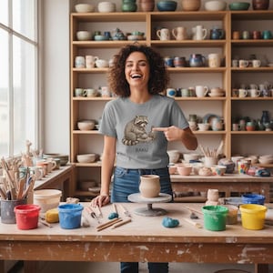 May include: A woman wearing a gray t-shirt with a raccoon graphic and the word "RAKU" in brown. She is smiling and pointing at a small ceramic pot on a pottery wheel. The background includes pottery shelves and art supplies.