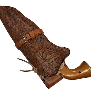 Könnte beinhalten: Eine braune Lederholster mit floralem und geometrischem Muster, die einen Revolver hält. Die Holster hat einen Schnallenverschluss und ein dekoratives Blumendesign. Der Revolver hat einen Holzgriff.