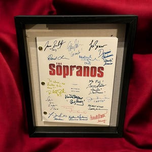 Pode incluir: Roteiro emoldurado da série de TV "Os Sopranos", com o título em vermelho e várias assinaturas. O roteiro é montado em um fundo bege dentro de uma moldura preta, em um fundo vermelho.