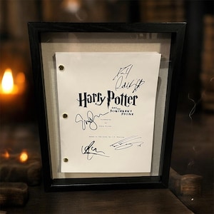 May include: Framed Harry Potter and the Sorcerer's Stone screenplay with signatures. The black frame surrounds a white paper with the title in a stylized font. The signatures are in black ink, and the paper is held in place with brass fasteners.