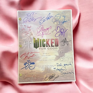 Puede incluir: Un programa firmado de "Wicked" con el título "Wicked For Good" en verde y rojo. El programa presenta firmas en varios colores, incluyendo azul, morado y rojo. El fondo muestra dos personajes del musical.
