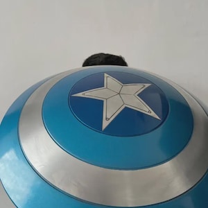 May include: A blue and silver shield featuring a white star emblem. The shield has a circular design with a blue outer ring and a silver band. The shield is a prop or costume accessory, likely for cosplay or themed events.