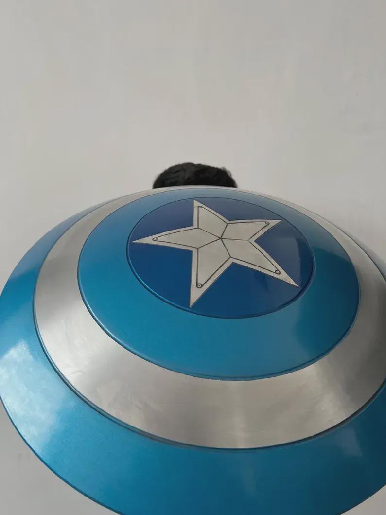 May include: A blue and silver shield featuring a white star emblem. The shield has a circular design with a blue outer ring and a silver band. The shield is a prop or costume accessory, likely for cosplay or themed events.