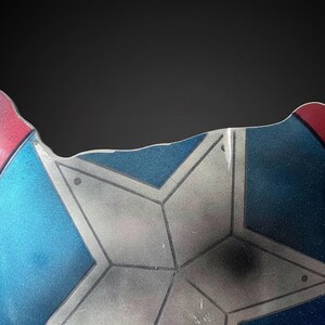 Puede incluir: Fragmento de un escudo con un dise&ntilde;o de estrella. El escudo presenta una combinaci&oacute;n de colores rojo, blanco y azul. El escudo tiene una apariencia desgastada, con desgaste visible, lo que sugiere una est&eacute;tica de batalla.