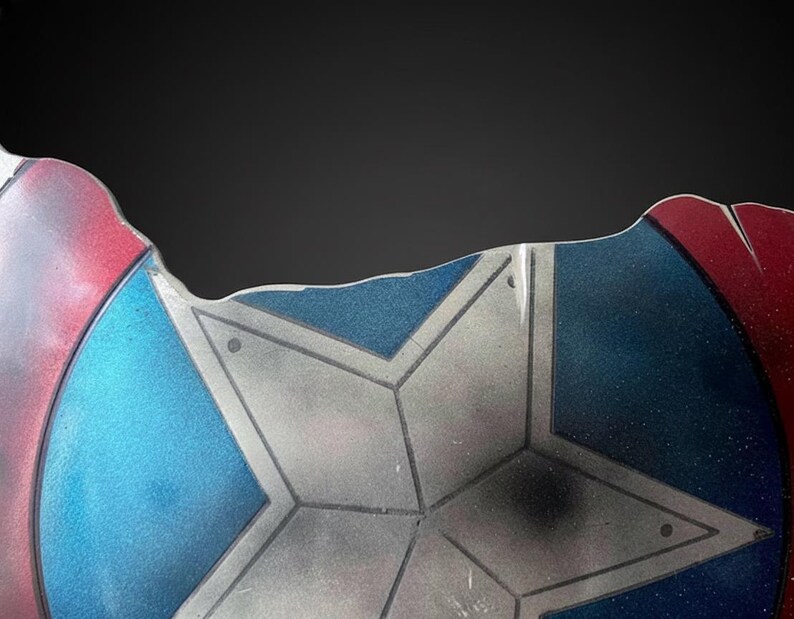 Puede incluir: Fragmento de un escudo con un dise&ntilde;o de estrella. El escudo presenta una combinaci&oacute;n de colores rojo, blanco y azul. El escudo tiene una apariencia desgastada, con desgaste visible, lo que sugiere una est&eacute;tica de batalla.
