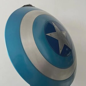 May include: A blue, silver, and white shield with a star emblem. The shield is round with concentric circles of colour. The central star is silver. The shield is a prop or costume accessory.