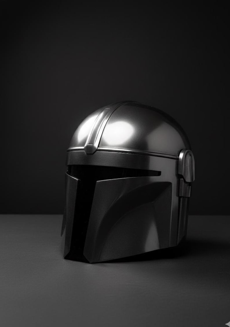 May include: A silver metallic helmet with a T-shaped visor and a rounded top. The helmet has a sleek, futuristic design, set against a dark background. It is likely a prop or collectible item.