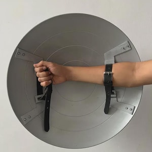 May include: A silver-coloured, circular shield with black leather straps and metal brackets. The shield has concentric circular ridges and is designed to be worn on the arm. The straps are secured with metal rivets.