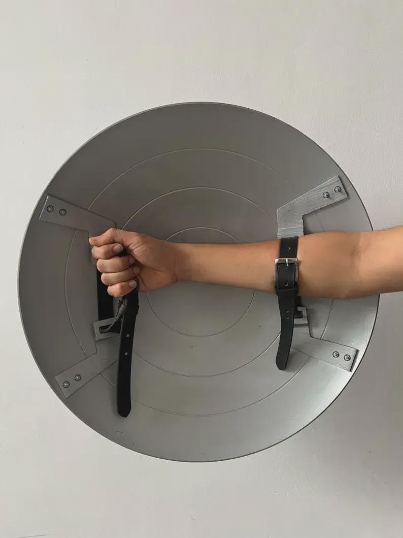 May include: A silver-coloured, circular shield with black leather straps and metal brackets. The shield has concentric circular ridges and is designed to be worn on the arm. The straps are secured with metal rivets.