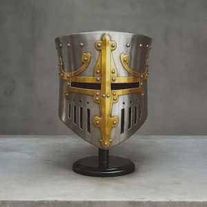 May include: A silver-colored medieval helmet with a gold cross design on the front. The helmet has rivets and vertical slits for vision and ventilation. It is displayed on a black stand against a gray background.