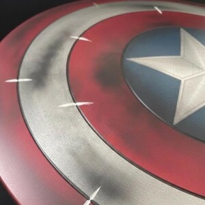 May include: A close-up of a Captain America shield. The shield features a red, white, and blue color scheme with a white star in the center. The shield has a distressed, metallic finish with white accent lines.