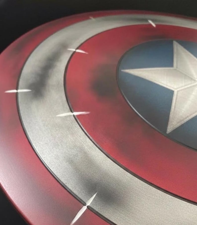 May include: A close-up of a Captain America shield. The shield features a red, white, and blue color scheme with a white star in the center. The shield has a distressed, metallic finish with white accent lines.