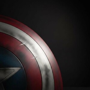May include: A close-up of a Captain America shield. The shield features a red and silver striped design with a blue star emblem. The shield has a distressed, battle-worn appearance, with visible scratches and wear.